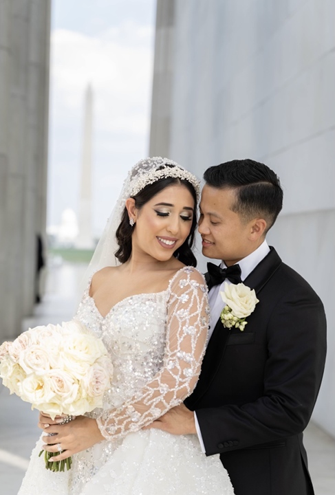A beautiful couple on their wedding day, embracing lovingly, with the groom in a black tuxedo and the bride in a white wedding gown holding a bouquet of white roses.